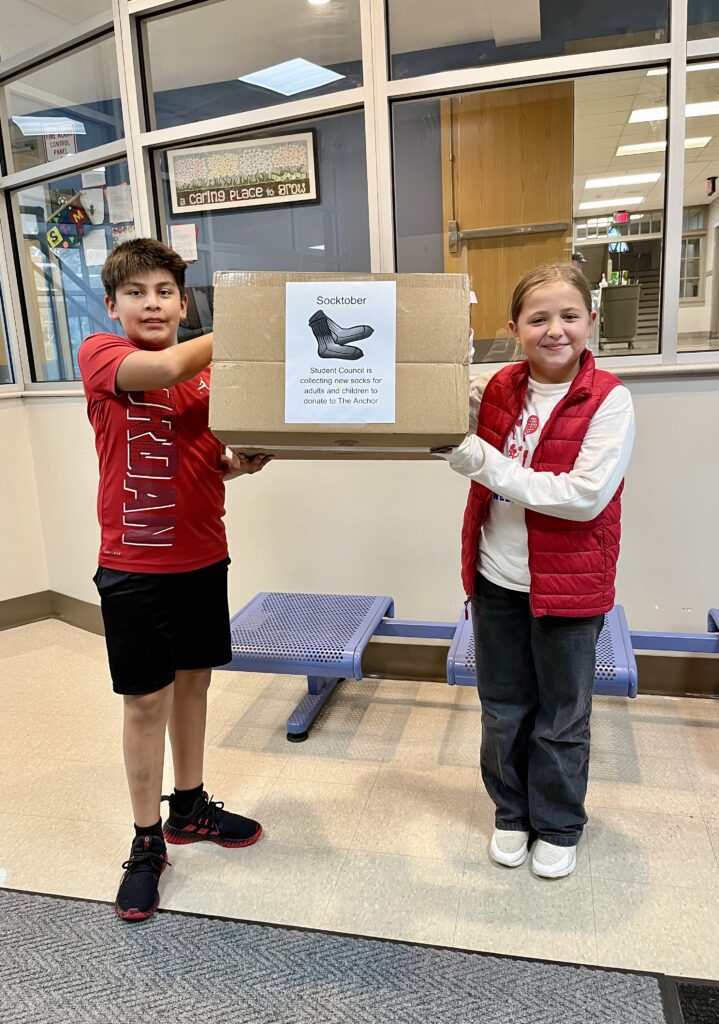 Student council members in elementary school lobby. Students are holding sock donation drive box.