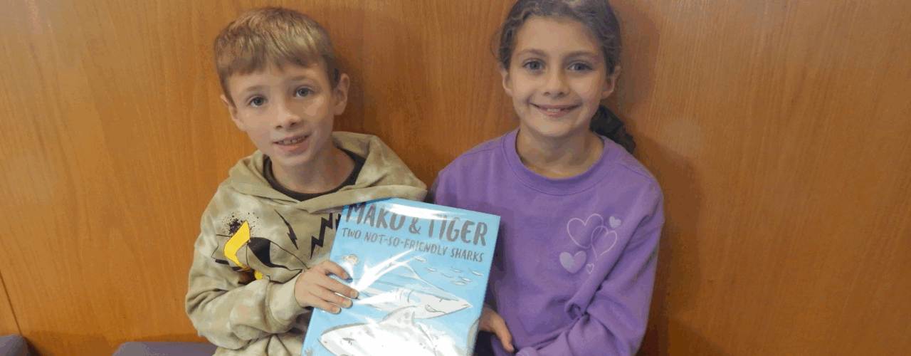A boy and a girl sitting together against a wood wall, smiling and holding a book.