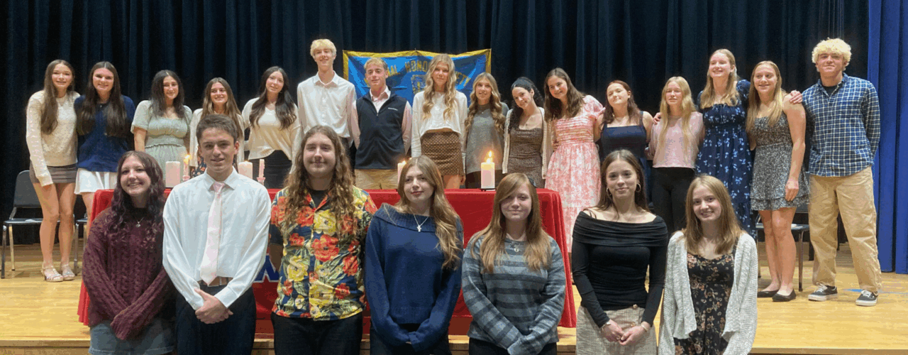 A large group of high school students in semi-formal attire posing for a photo on a stage. They stand behind a red-clothed table featuring lit candles, with a blue "National Honor Society" banner hanging in the background.