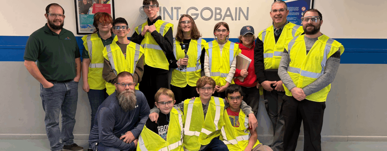 A group of students and three adults posing for a photo in front of a "SAINT-GOBAIN" logo on a wall.