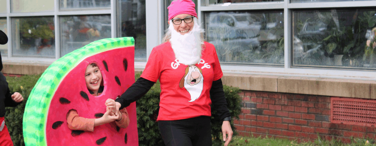 An adult dressed as the dwarf "Grumpy," holding hands with a student in a large watermelon slice costume.