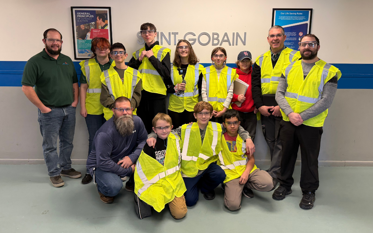 A group of students and three adults posing for a photo in front of a "SAINT-GOBAIN" logo on a wall.