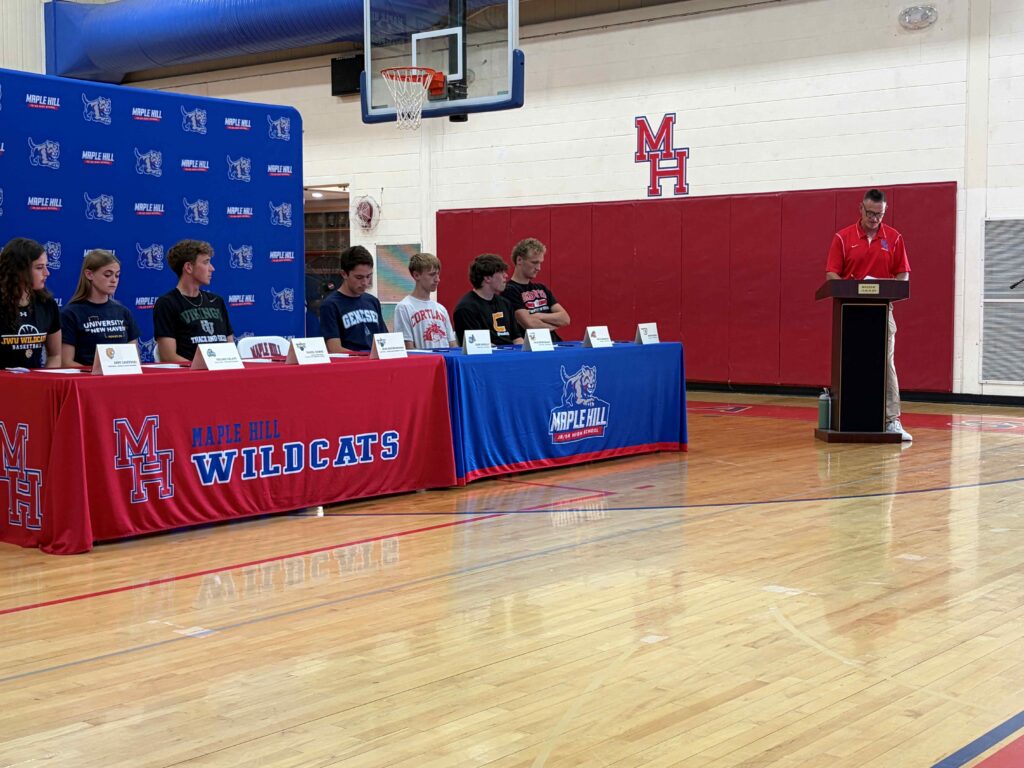 A group photograph of eight student-athletes seated side-by-side at a long table during a college athletics signing ceremony at Maple Hill High School.