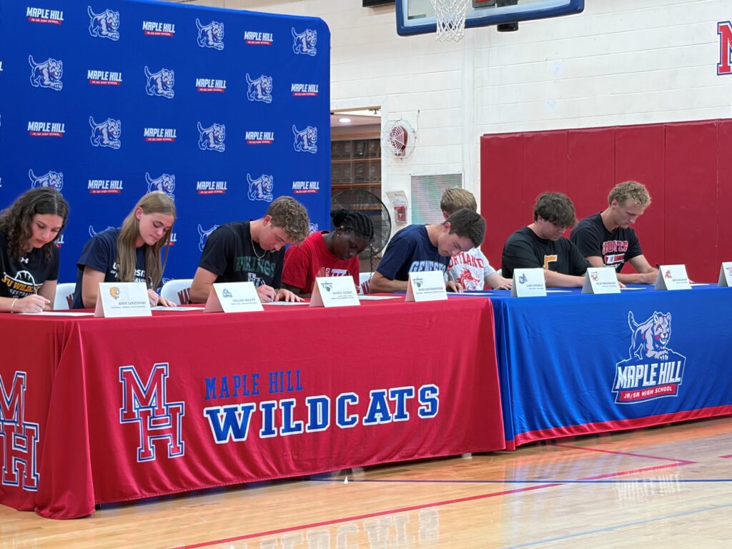 A group photograph of eight student-athletes seated side-by-side at a long table during a college athletics signing ceremony at Maple Hill High School.