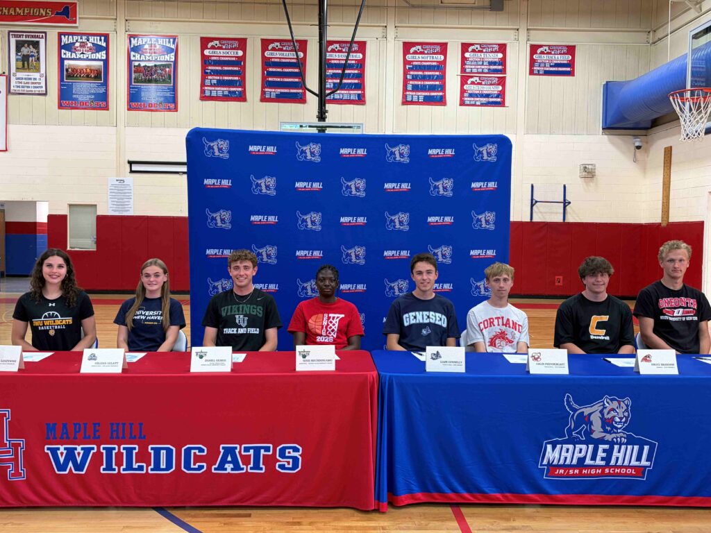 A group photograph of eight student-athletes seated side-by-side at a long table during a college athletics signing ceremony at Maple Hill High School.