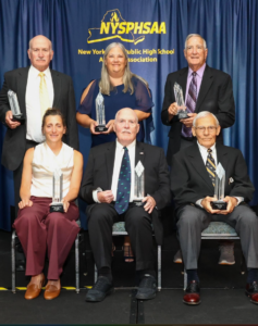 Group photo of the NYSPHSAA Hall of Fame inductees standing and seated on stage, holding crystal awards in front of a blue backdrop.