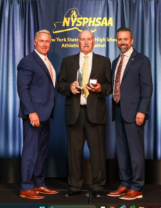 Coach Dan Gillespie stands with two officials holding his NYSPHSAA Hall of Fame crystal trophy and commemorative ring box.