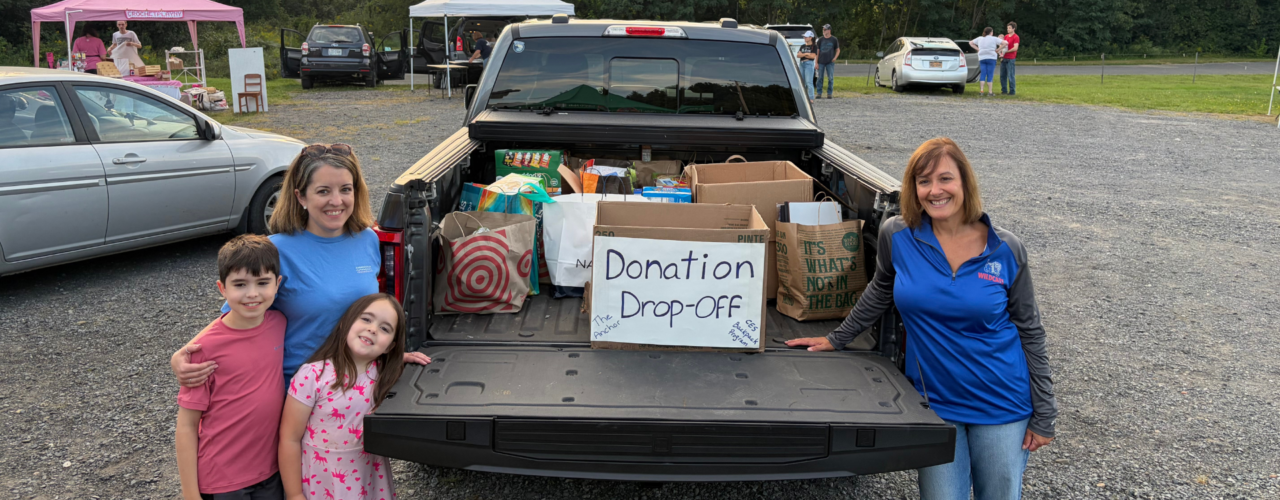 Elementary school staff standing by an open pickup truck bed loaded with donated items next to a 'Donation Drop-Off' sign.