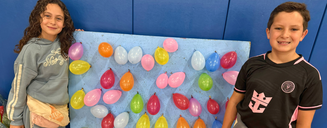 Two students posing beside a board covered in colorful balloons set up for a carnival-style balloon pop game.