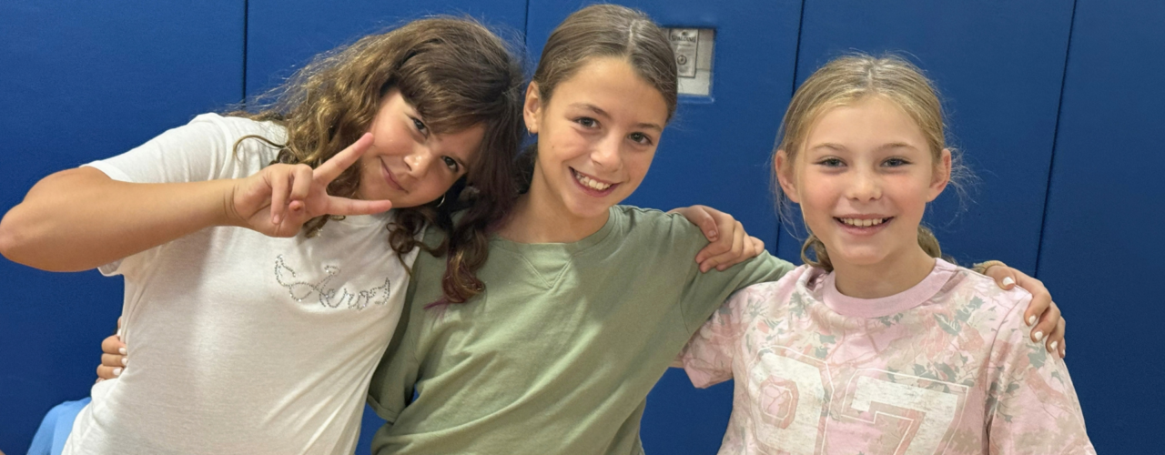 Three young girls posing together with arms around each other, with one flashing a peace sign.